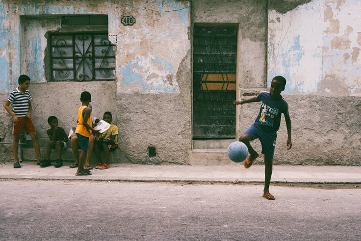 This may contain: a young man kicking a soccer ball on the street while people watch from the sidewalk
