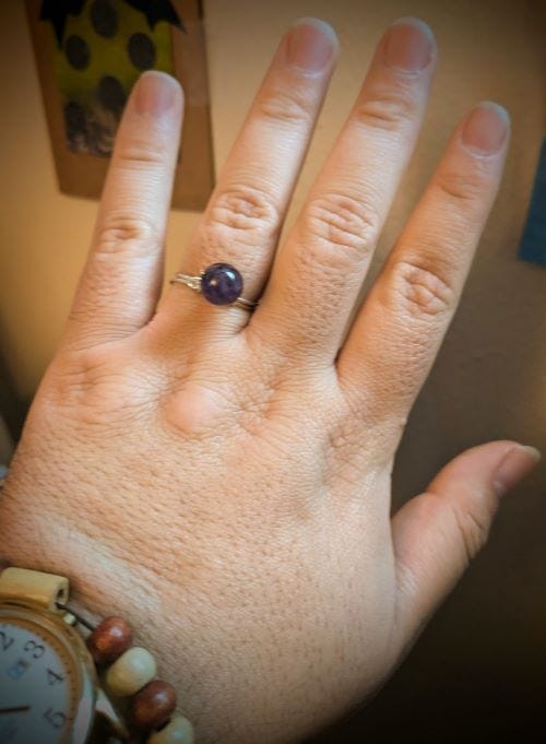 hand with the back facing the camera, showing a metal ring with a purple bead on top