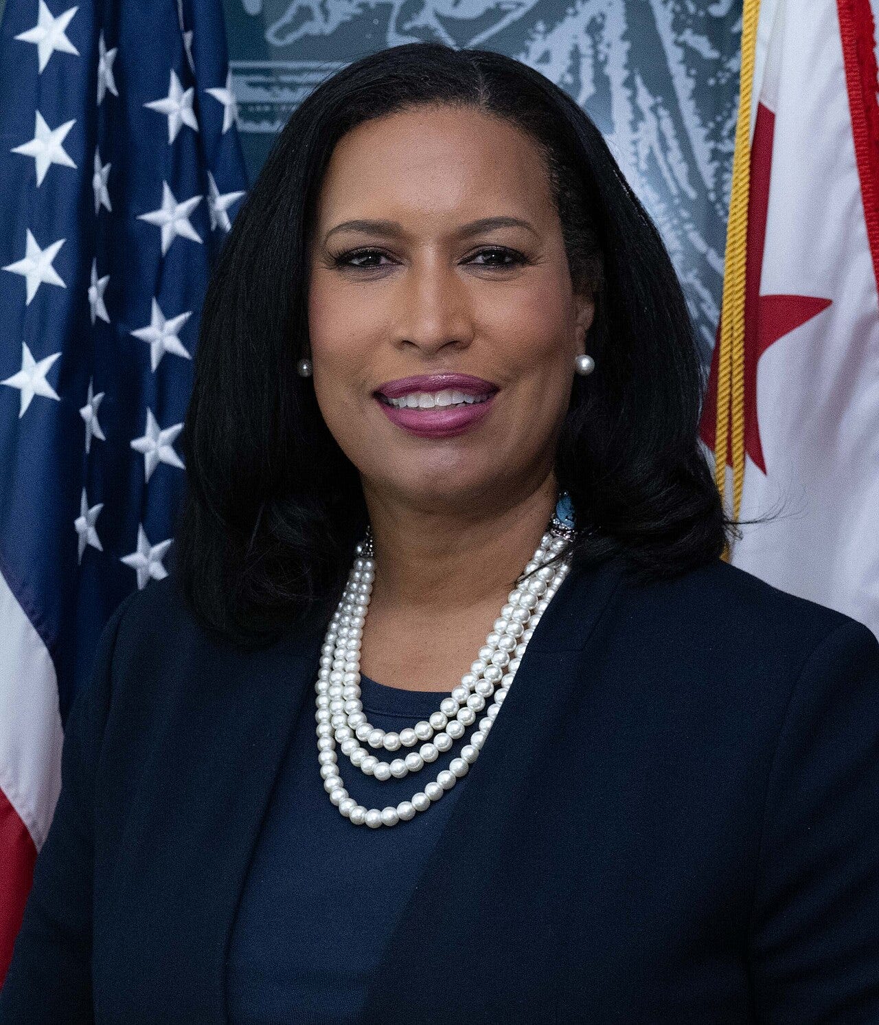 official portrait of Muriel Bowser; mayor of DC. straight black hair; dark suit, pearls, flag in background