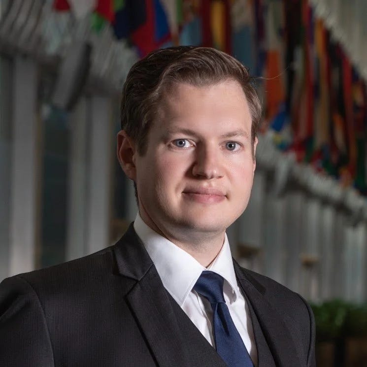 Headshot of in a dark suit and navy tie standing before a row of international flags.