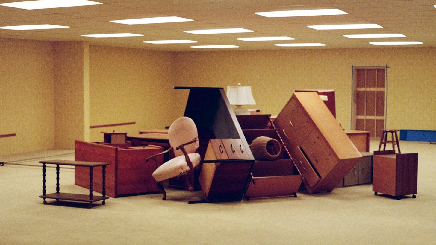 still from backrooms; an office room with low ceilings and flourescent lights in which a bunch of furniture has been stacked haphazardly in the middle of the room
