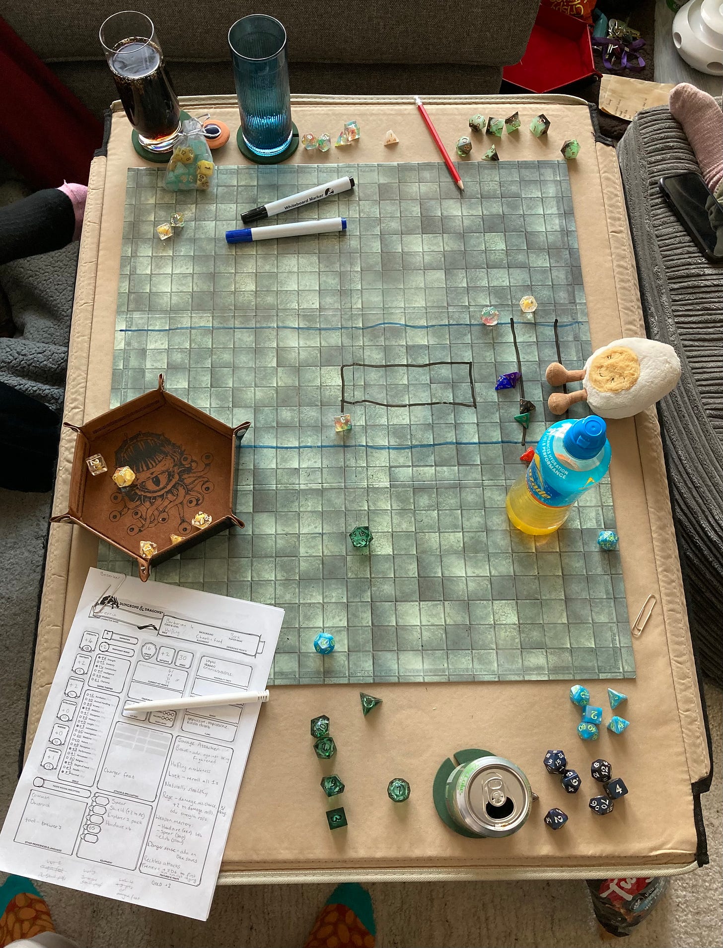 Photo of a dungeons and dragons game, with dice scattered over the grid, several drinks around the outside, and dippy the soft toy egg sitting at the top watching over the chaos