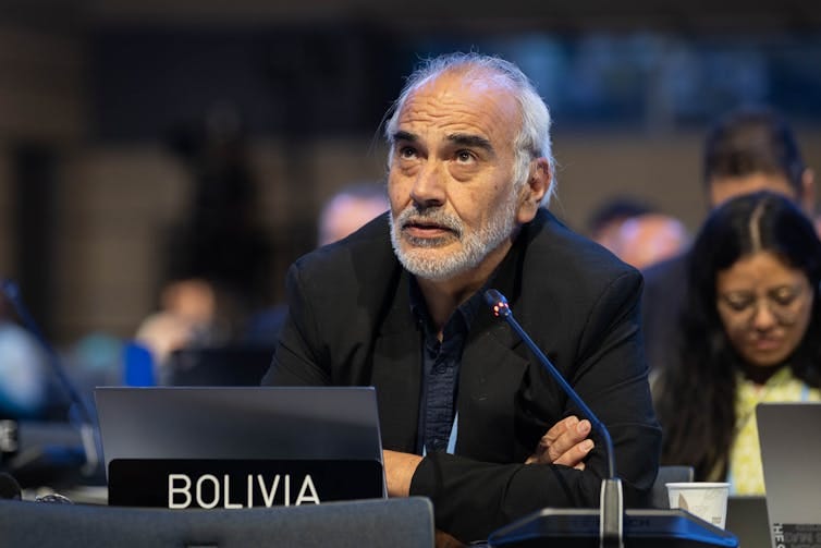 A man with his arms crossed leans on a desk with a 'Bolivia' label in front of it.