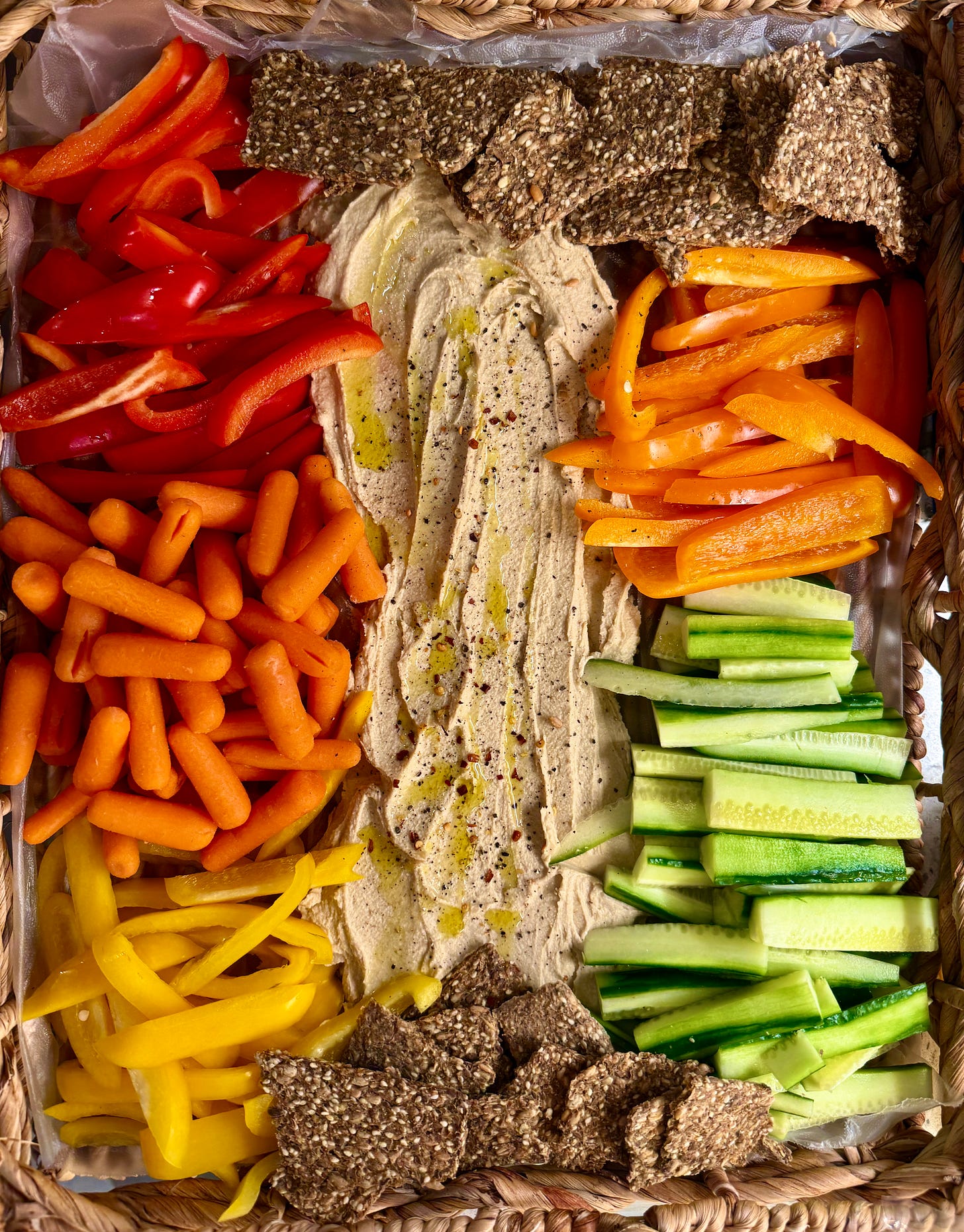 A platter of fresh vegetables and snacks arranged around a center of garlic hummus, including red, yellow, and orange bell pepper slices, English cucumber slices, baby carrots, and gluten-free seed crackers, with the hummus spread smoothly in the middle and garnished with olive oil, red pepper flakes, and black pepper. A platter of fresh vegetables and snacks arranged around a center of garlic hummus, including red, yellow, and orange bell pepper slices, English cucumber slices, baby carrots, and gluten-free seed crackers, with the hummus spread smoothly in the middle and garnished with olive oil, red pepper flakes, and black pepper.