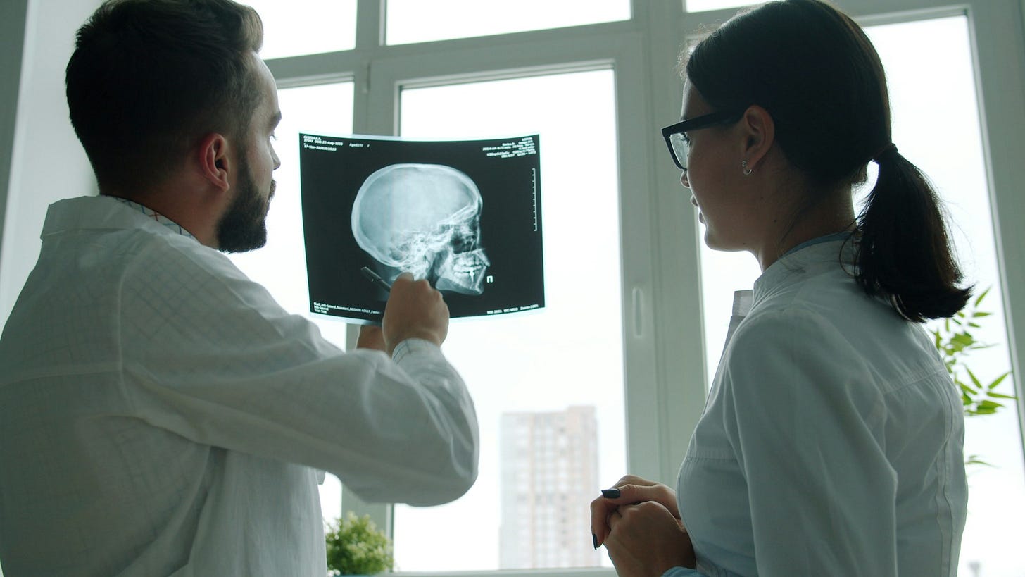 Two healthcare professionals study a skull X-ray near a bright clinic window, highlighting the role of teaching and clinical guidance in medical training. Photo by Vitaly Gariev on Unsplash
