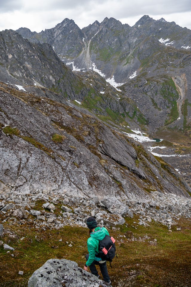 A hiker in a green rain jacket, black beanie, and black pack descends a slope of tundra and scattered boulders away from the camera. Far below, a small blue-green pond sits at the base of the valley, and the wall of peaks beyond rises dark and snow-streaked into overcast light. A hiker in a green rain jacket, black beanie, and black pack descends a slope of tundra and scattered boulders away from the camera. Far below, a small blue-green pond sits at the base of the valley, and the wall of peaks beyond rises dark and snow-streaked into overcast light.