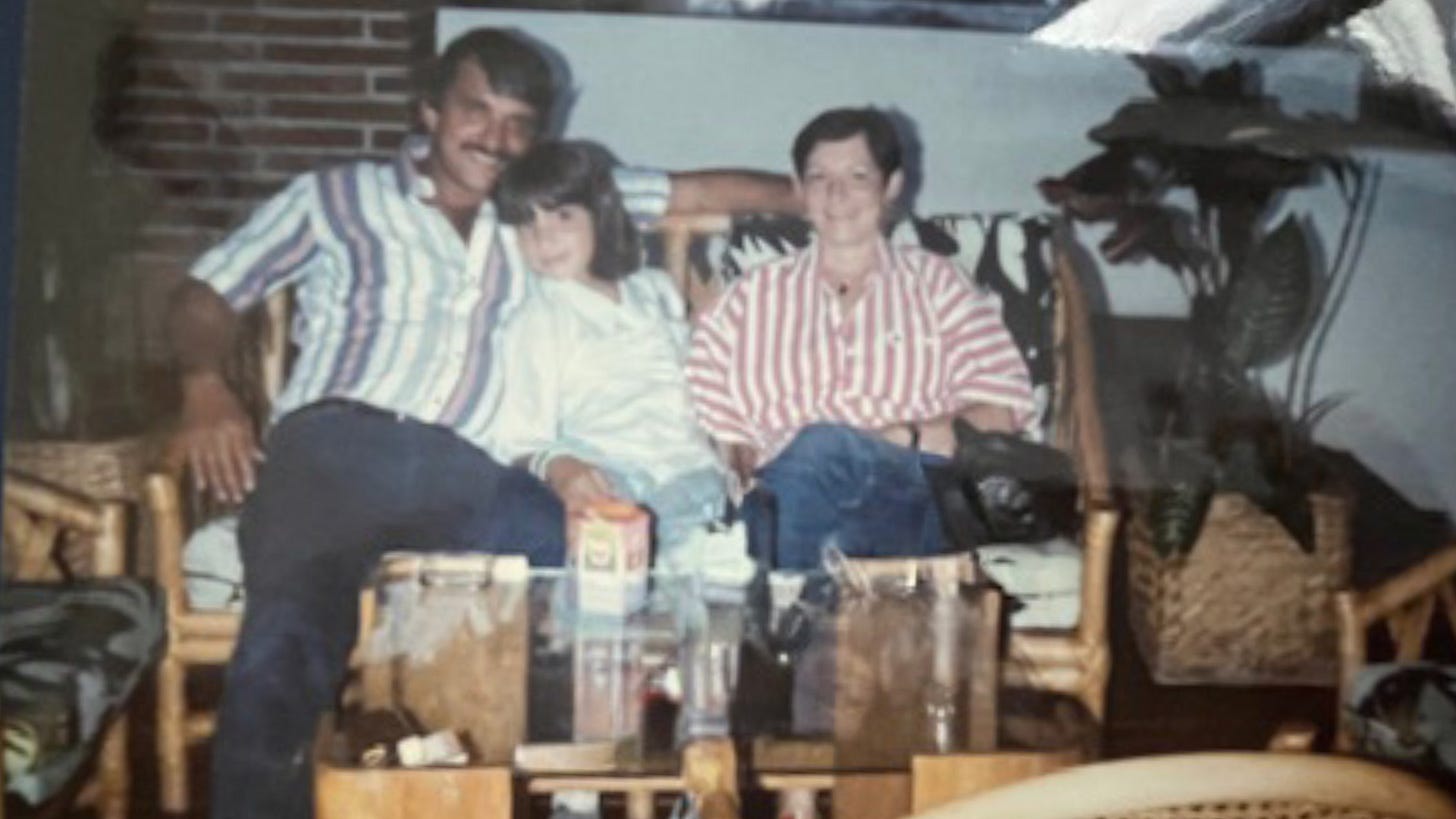 A photo of three people sitting closely together on a wicker couch in a cozy living room. An adult man on the left, a child in the middle, and an adult woman on the right all smile at the camera. There are drinks and a carton on the table in front of them, and plants and a brick wall in the background.