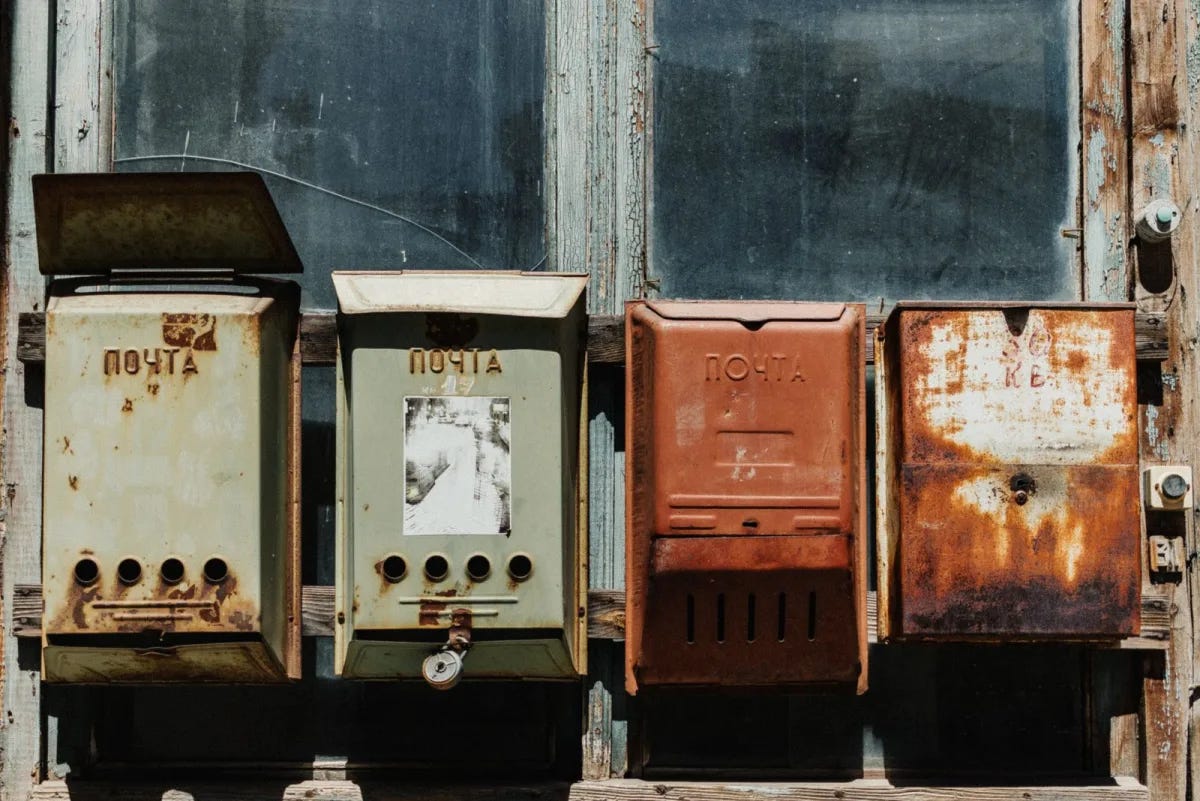 Four old mailboxes in a row on a weathered wooden wall, each a different color