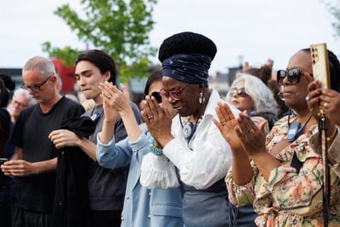 Craig, hands together as if in prayer, with people either side. 