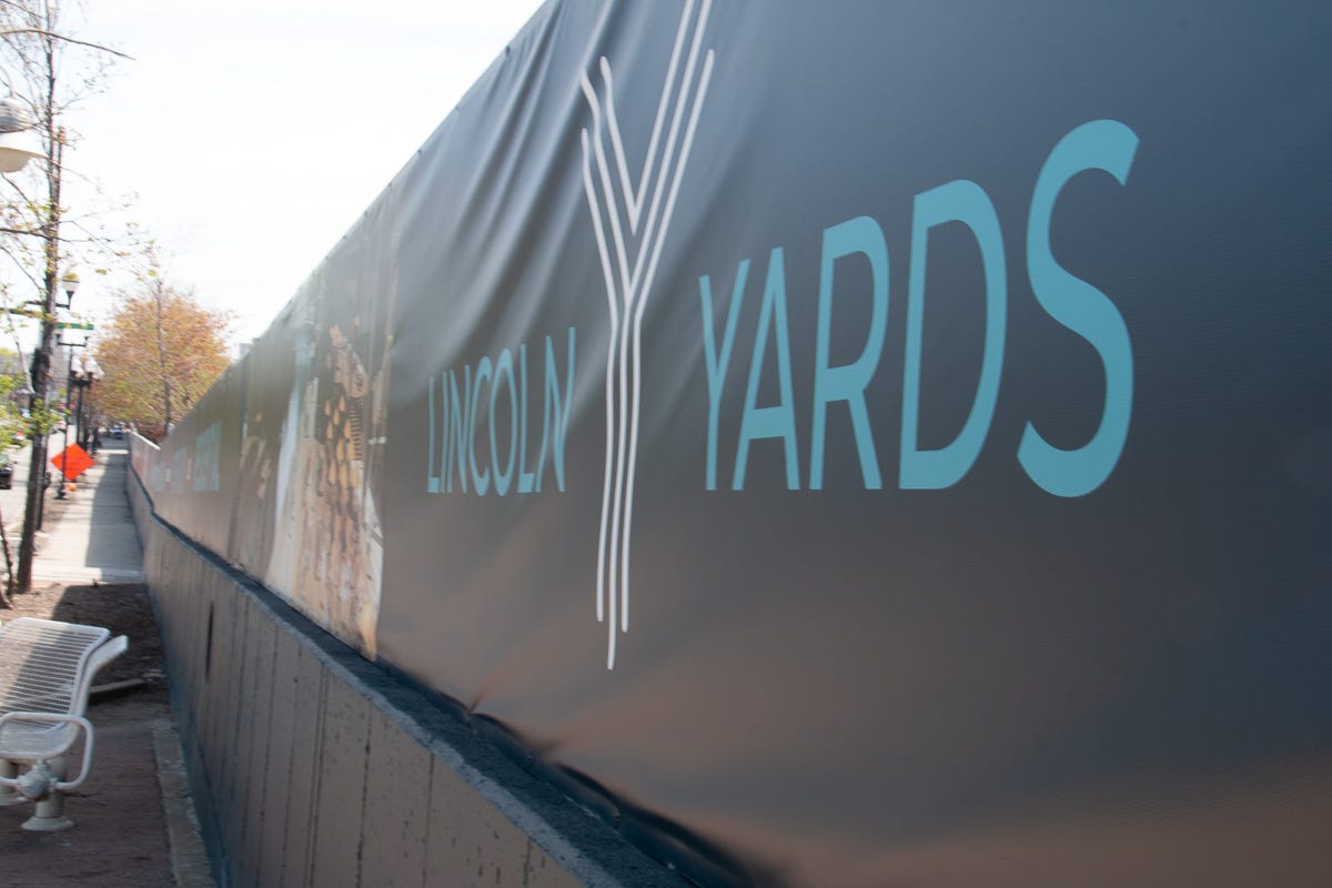 View down a sidewalk with a long black fence displaying the words “Lincoln Yards” in light blue letters. View down a sidewalk with a long black fence displaying the words “Lincoln Yards” in light blue letters.