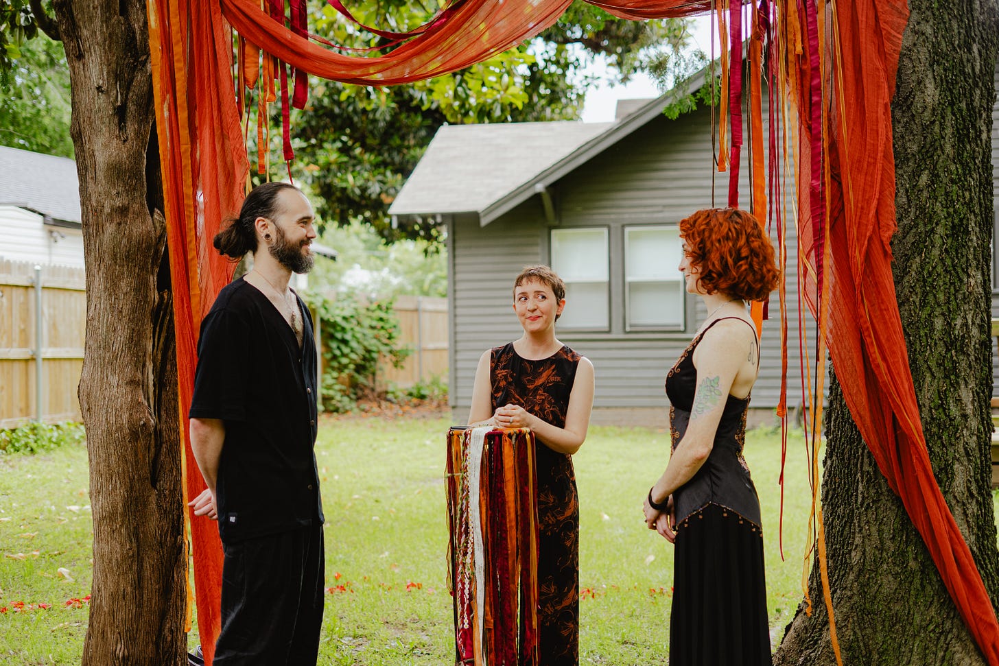 a backyard wedding: on the left, Jo's parter Taylor, on the right, Jo, and Maeg in the middle playfully peering sidelong at Jo, who giggles. they are between two trees which are draped in deep golen orange gauze and ribbons of gold and deep red/purple. 