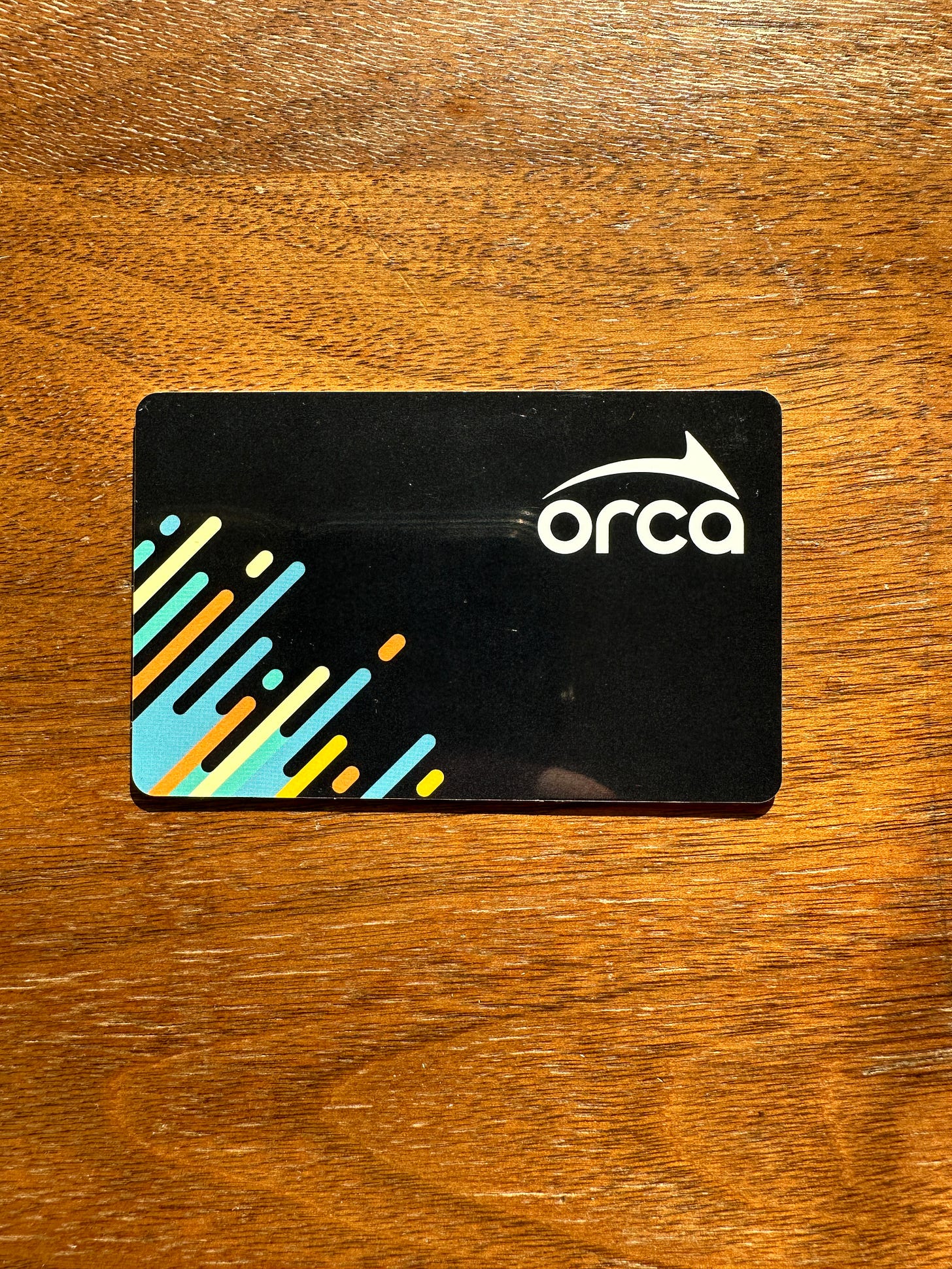 Black rectangular transit card with rounded corners. The ORCA logo is in the top right corner in white letters There are also some colorful stripes on the card. The card is sitting on a wooden table. Black rectangular transit card with rounded corners. The ORCA logo is in the top right corner in white letters There are also some colorful stripes on the card. The card is sitting on a wooden table.