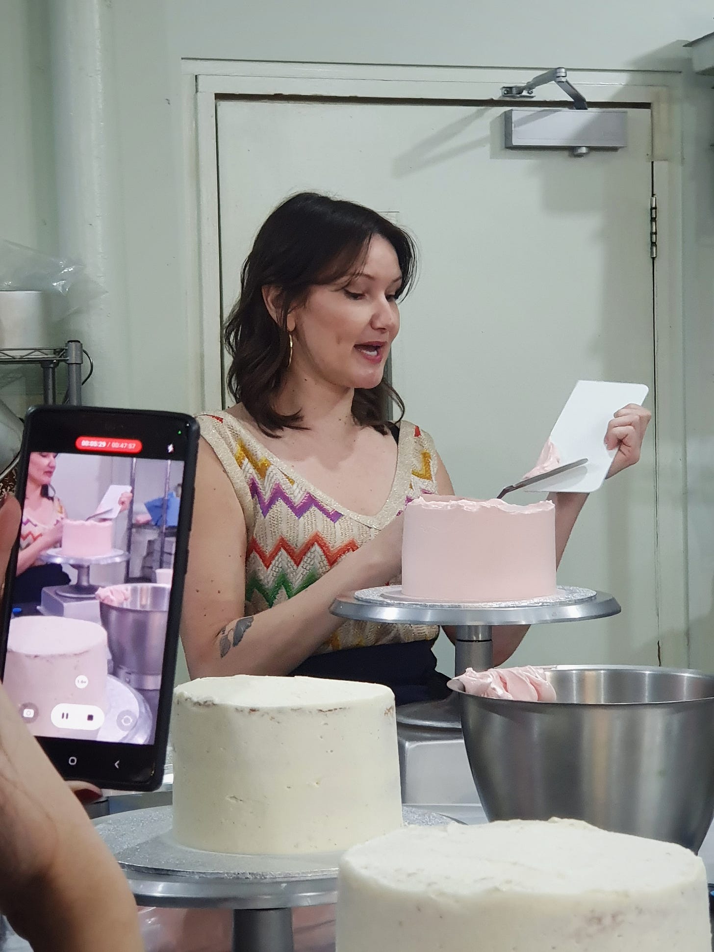 Photo of a woman talking while she ices a cake. There is another screen in the photo taking the same photo.