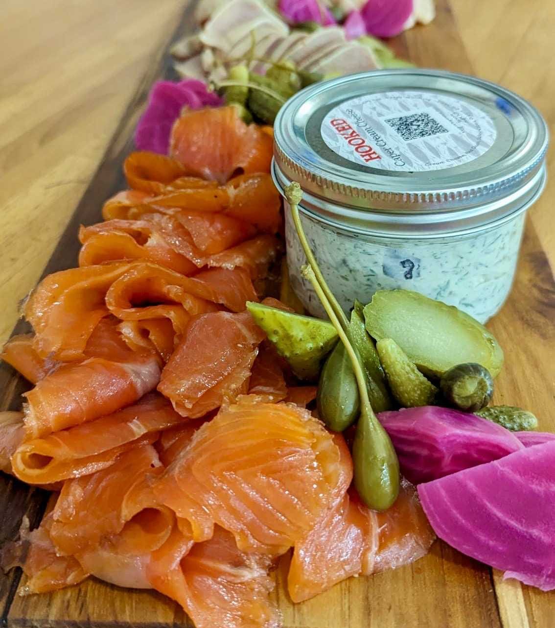 A photo of a long wood board on a wood counter covered with bright orange smoked salmon, deep green pickles and caperberries, pinkish pickled beets and a jar of Hooked Caper Cream Cheese