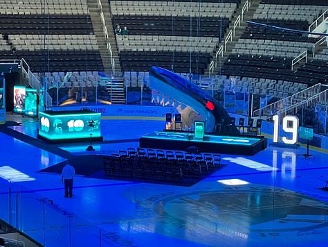 Photo 1: Image of woman in Sharks jersey smiling outside arena; Photo 2: Image of Joe Thornton ice sculpture; Photo 3: Image of Joe Thornton coming through Shark head on ice; Photo 4: image of two women smiling with ice rink behind them; Photo 5: Image of Shark head and chairs on ice for ceremony; Photo 6: Image of Joe Thornton on big screen; Photo 7: Image of Thank You Joe box where jersey was raised with family behind it; Photo 8: Image of ice rink with Shark head and players; Photo 9: Image of #12 and #19 jersey's hanging in the rafters of the arena