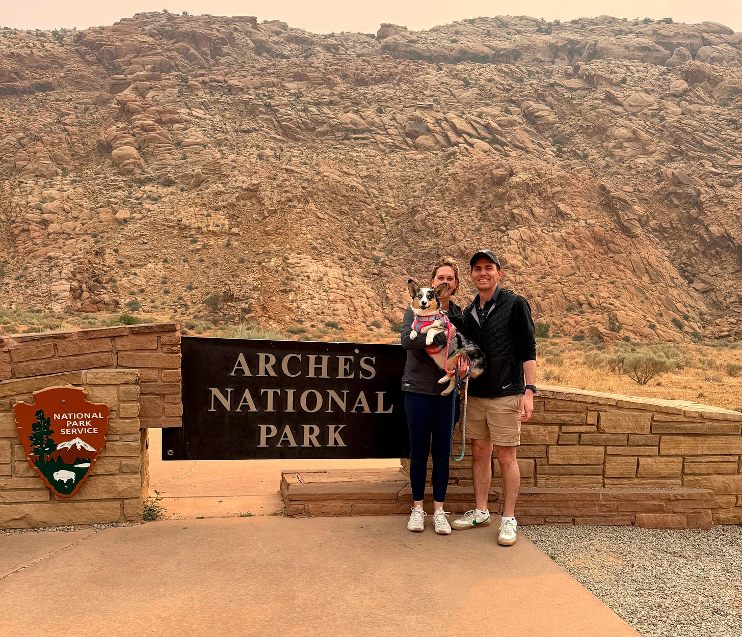 Kim and her husband Joe stand in front of the sign for Arches National Park with the National Park Service logo off to the side. Kim holds her blue merle corgi who is wearing a pink harness. The sky is hazy from smoke.