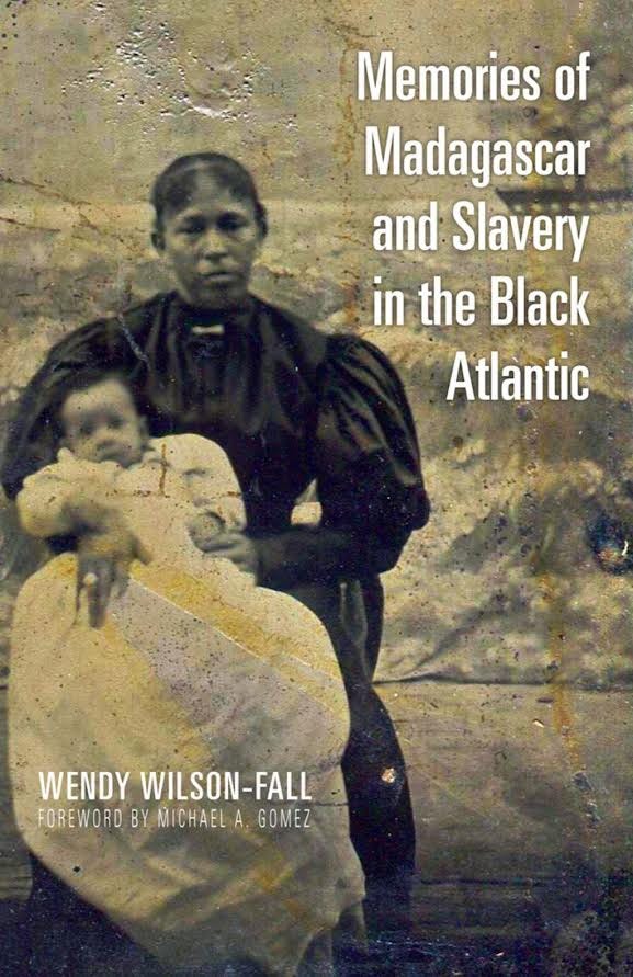 Pictured is a Malagasy enslaved woman holding a baby who is covered in an oversized white blanket and a white dress underneath. She is resting this baby across her legs, and a large sailboat is placed behind her on a coastline in the top right corner in the background of the cover with the title “Memories of Madagascar and Slavery in the Black Atlantic” indented over the boat in large white font.
