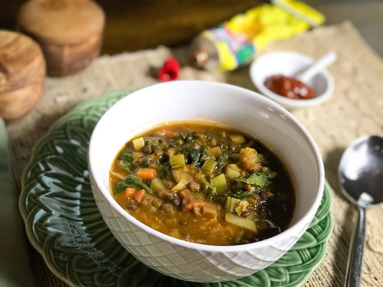 Sunday Souper Soup with lentils, carrots, celery, onions and greens in a white bowl on a green Tiffany basket-weave plate. In the back ground is harissa from a tube.