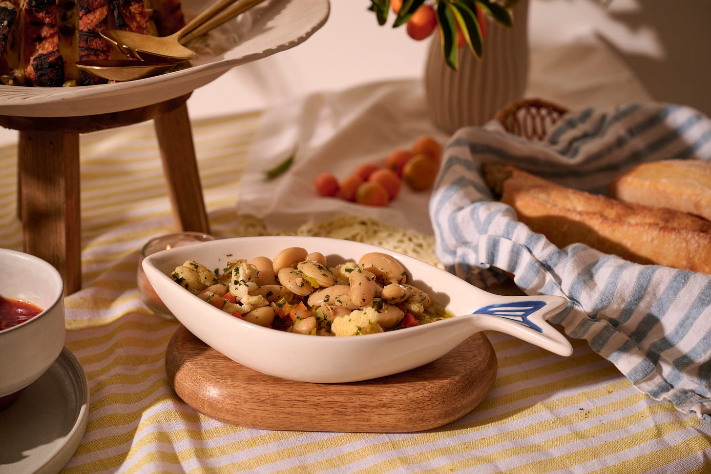 Marinated gigante beans with cauliflower and peppers in a fish-shaped dish, served alongside grilled spiedini, fresh baguettes, and apricots on a sunlit summer table.