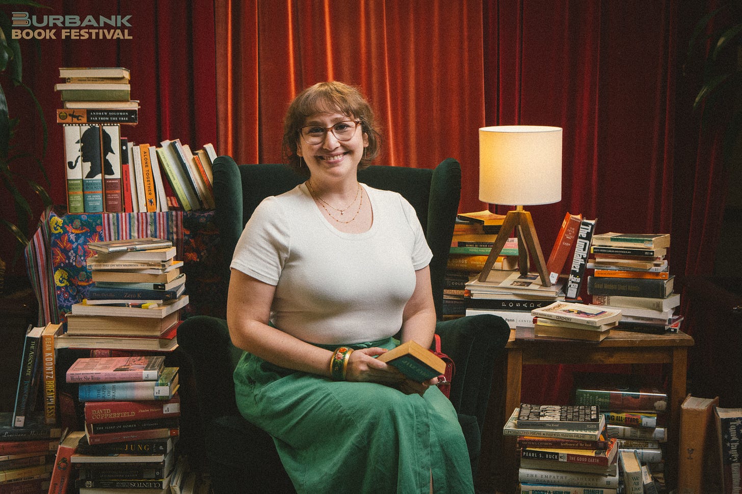 Kat Spada sits on a dark green velvet chair in front of a red curtain. Stacks of books are piled all around her. She wears a white tee shirt, green skirt, glasses, and a smile. The logo for the Burbank Book Festival watermarks the image.