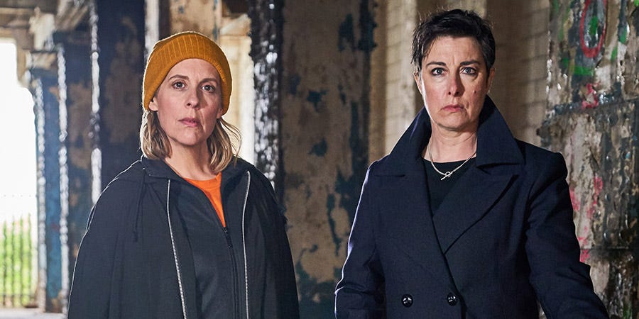 In an outdoor industrial space, two white women stand facing forward. The blonde woman on the left wears a black jacket and a mustard-colored beanie; she has a serious look on her face as she looks into the camera. The taller woman with short dark hair is wearing a black peacoat and is staring seriously into the camera. 