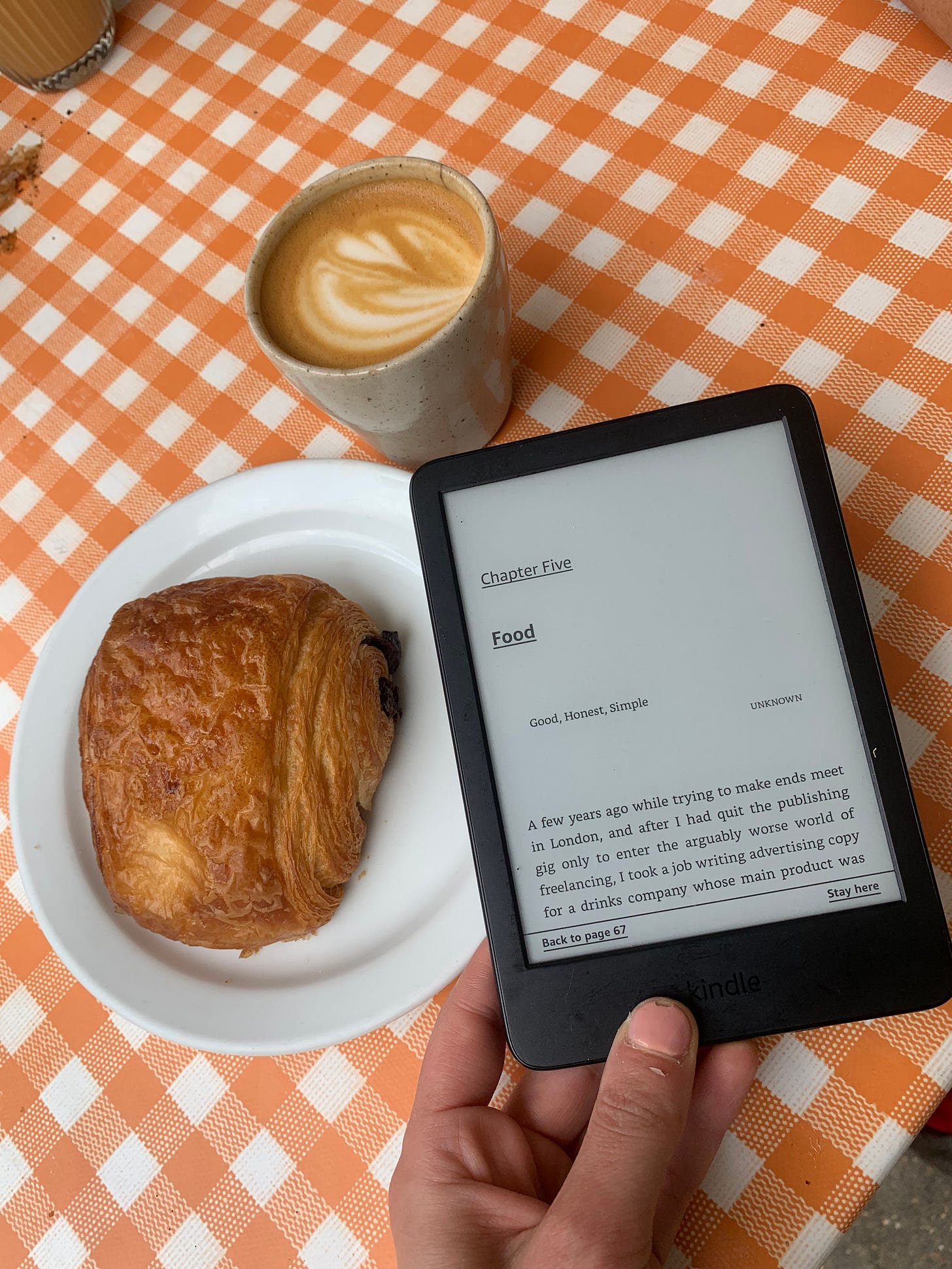 A hand holding an e-book version of Nathalie Olah's 'Bad Taste' with the chapter titles, 'Food' showing on the front screen. A coffee and a french pastry sit in the background.