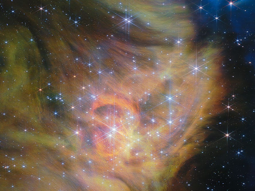 A star-forming region. The left two thirds of the image is blanketed in thick clouds of green and yellow gas and dust that form swirling patterns. Some of the gas is spread into thin, thready wisps. The right third of the image is the black background speckled with stars and multi-colored dots that represent distant galaxies. Bright stars are scattered throughout the clouds, with the densest cluster situated in the center of the scene. Each is crowned with six long spikes and two shorter spikes created by the optics of the telescope’s mirrors. In the upper right, a couple of hidden stars blast out long, glowing jets of material that glow in blue.