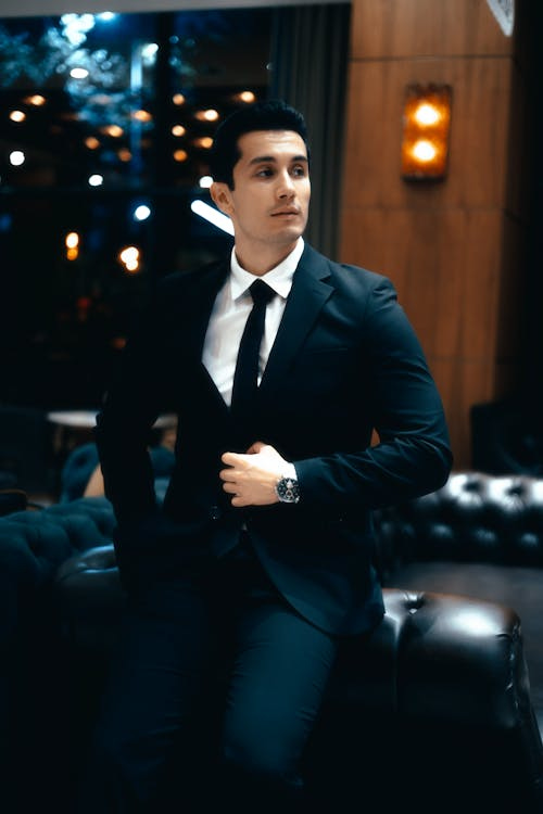 Free A confident young man in a refined suit seated in an upscale lounge setting. Stock Photo