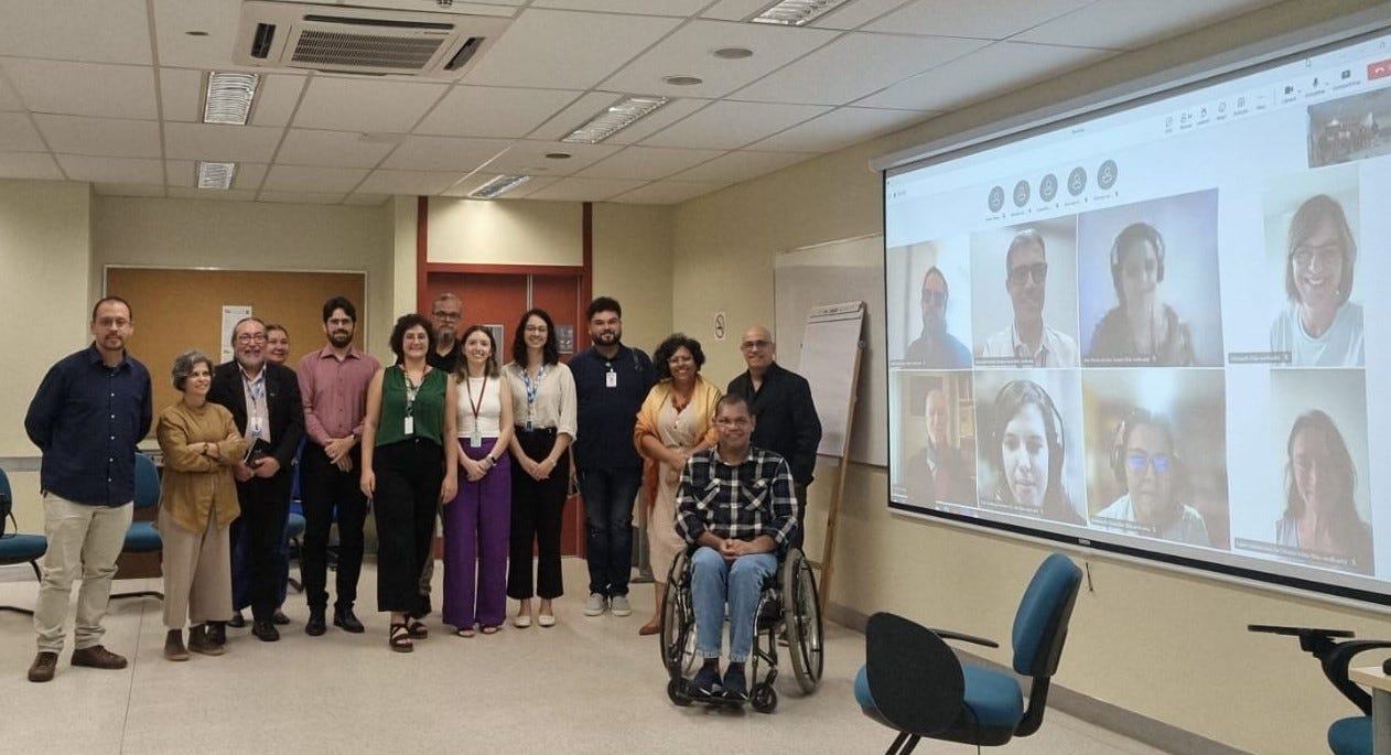 Descrição: Em uma sala de reuniões, um grupo posa para a fotografia. À esquerda, um outro grupo e, à direita, uma tela na qual estão projetadas oito pessoas em ambiente virtual. Todos os grupos são formados por homens e mulheres de diferentes etnias e idades.