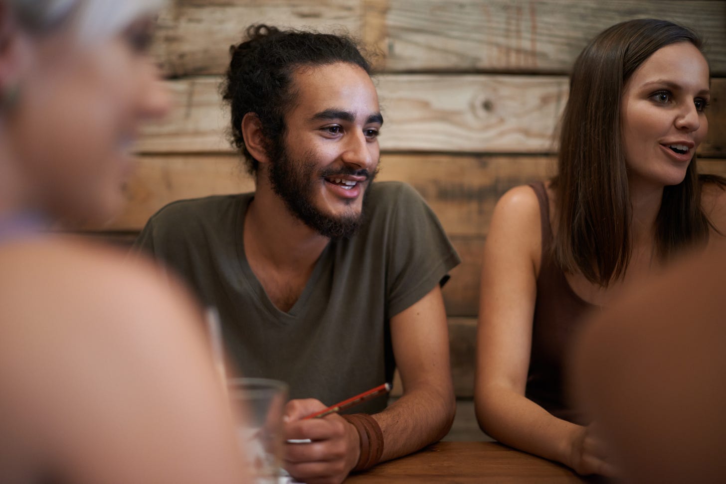 A young man with a beard and tied-back curly hair smiles while seated at a wooden table next to a woman with straight brown hair. They appear to be in friendly conversation, with blurred figures in the foreground suggesting a group setting. A young man with a beard and tied-back curly hair smiles while seated at a wooden table next to a woman with straight brown hair. They appear to be in friendly conversation, with blurred figures in the foreground suggesting a group setting.