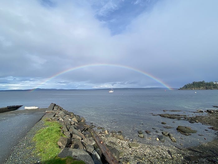 One sailboat sits under a perfect rainbow over the ater. Green moss is on the shore.