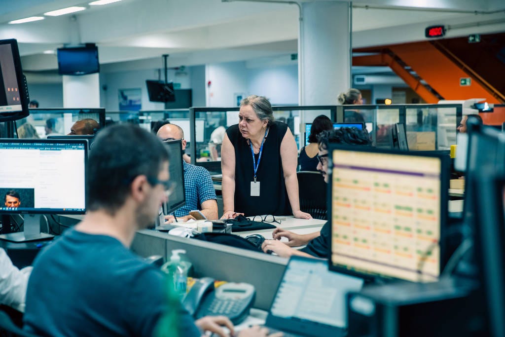 A imagem mostra uma redação de jornal com várias pessoas trabalhando em estações de trabalho. No primeiro plano, um homem de camiseta escura está concentrado em seu computador, enquanto uma mulher de cabelo grisalho e blusa preta está em pé, interagindo com os colegas. Ao fundo, há várias telas de computador exibindo informações e gráficos. O ambiente é bem iluminado e organizado, com divisórias entre as mesas. A imagem mostra uma redação de jornal com várias pessoas trabalhando em estações de trabalho. No primeiro plano, um homem de camiseta escura está concentrado em seu computador, enquanto uma mulher de cabelo grisalho e blusa preta está em pé, interagindo com os colegas. Ao fundo, há várias telas de computador exibindo informações e gráficos. O ambiente é bem iluminado e organizado, com divisórias entre as mesas.