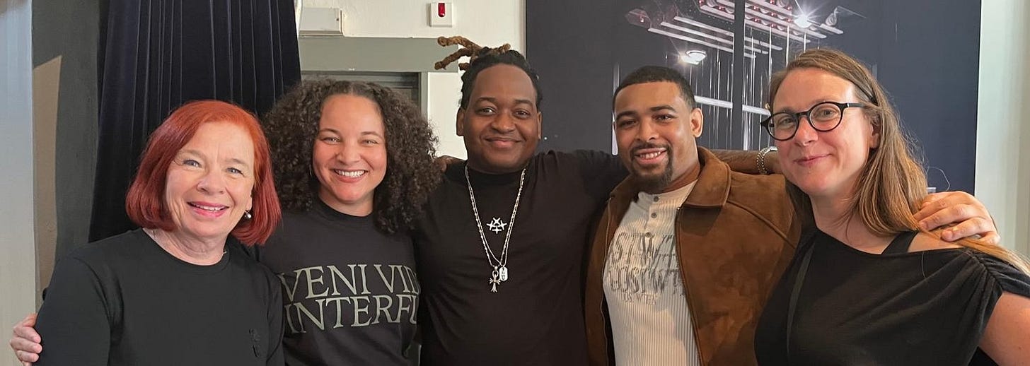 Five people pose together with arms around each other, smiling at the camera. From left to right: a woman with short red hair in a black top, a woman with curly hair wearing a black "Veni Vidi Interfui" shirt, a man with locs wearing a black turtleneck and chain necklaces, a man in a brown suede jacket over a graphic shirt, and a woman with shoulder-length hair and glasses in a black top. A dark backdrop with a photograph is visible behind them.