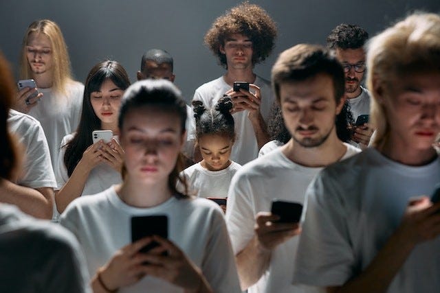 dim room full of people dressed in white t-shirts. they are facing the camera and looking down at phones in their hands