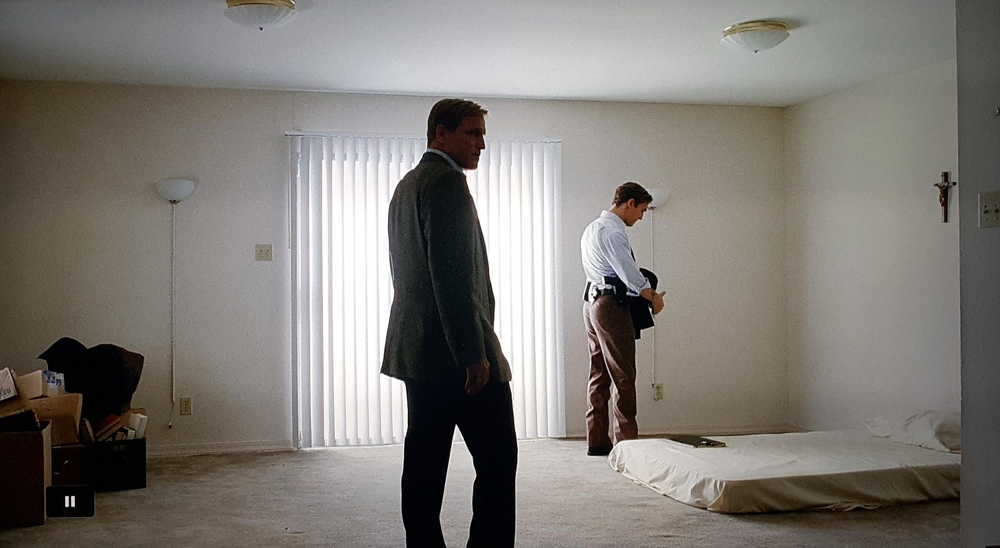 Two suited detectives stand in a bare apartment living room. There's a mattress on the floor and a crucifix above it.