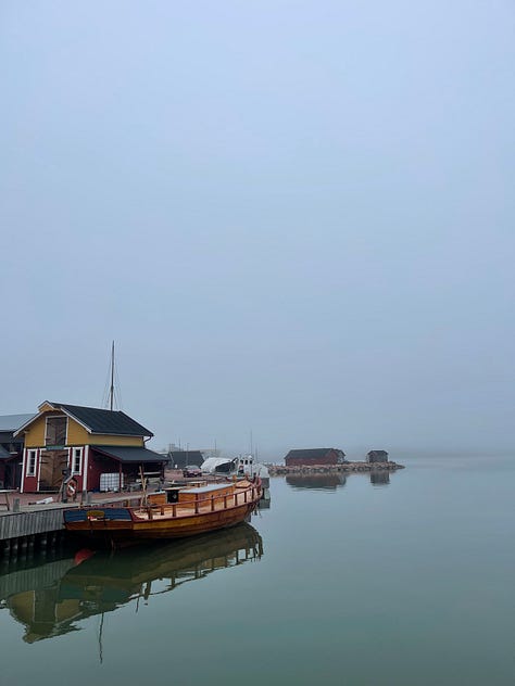 A blue sky with clouds and sky reflected in the water, a foggy day with grey sky, grey, still water and a half built wooden boat in the water moored to the pier, 3: looking out to water and fog with wires from rigging spanning the photo corners and the end gear of the deck railing in the foreground.