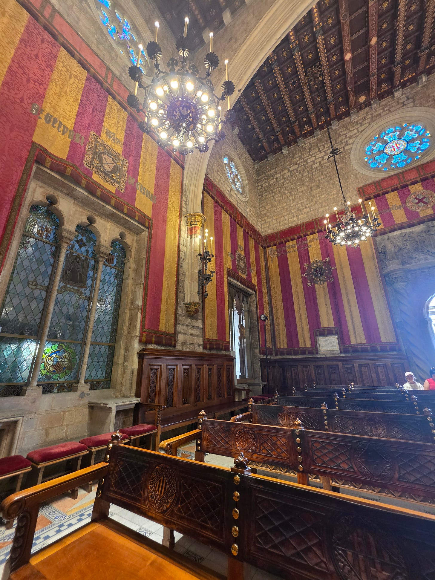Interior del Salón del Consell de Cent en el Ayuntamiento de Barcelona con tapices de rayas rojas y amarillas y ventanales góticos y techo de madera