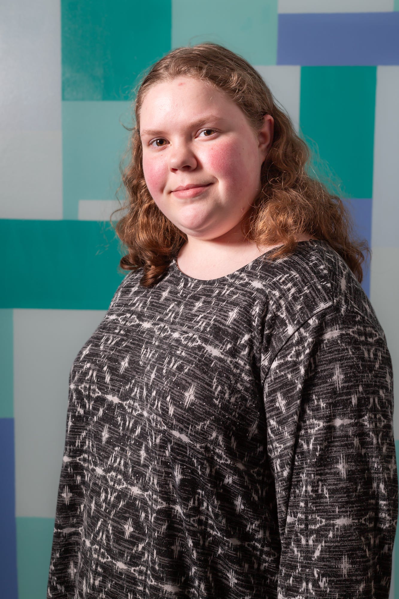 Portrait of a 15 year old white skinned woman. She has shoulder length blonde curly hair and light brown eyes. She has a slight smile. She is wearing a long sleeved black shirt with a white pattern.