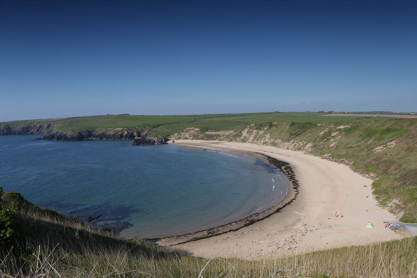 A high-angle view of a picturesque, crescent-shaped sandy beach on a bright, sunny day with a clear blue sky. Calm, blue water fills the bay, meeting pale sand that is bordered by rolling, grass-covered green cliffs. A dark line of washed-up seaweed traces the sweeping curve of the shoreline, and a few tiny figures of people can be seen enjoying the vast, quiet beach.