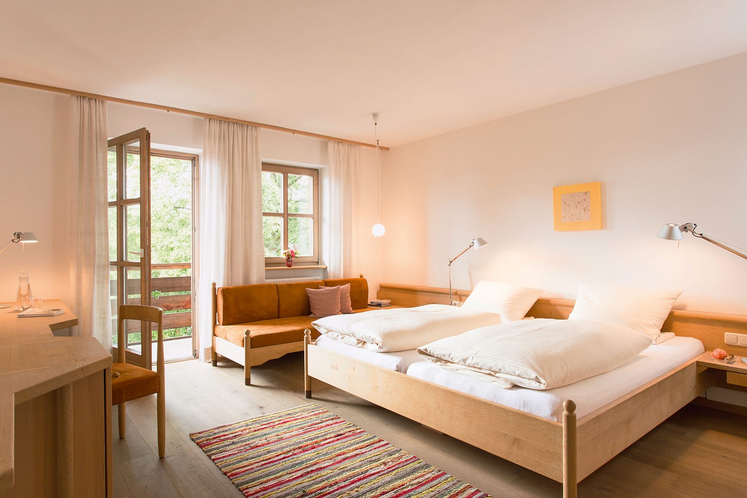 A bedroom at the Pausnhof with pale wooden furniture and white walls. It looks very calming and outside the windows are leafy green trees.