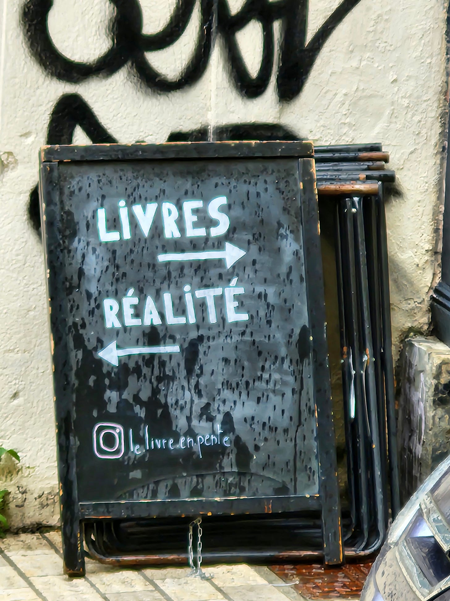 A black sandwich board stands propped against a white wall with graffiti on it. The sign says, in white block letters, "Livres" (with an arrow pointing right) and "Réalité" with an arrow pointing left. A black sandwich board stands propped against a white wall with graffiti on it. The sign says, in white block letters, "Livres" (with an arrow pointing right) and "Réalité" with an arrow pointing left.
