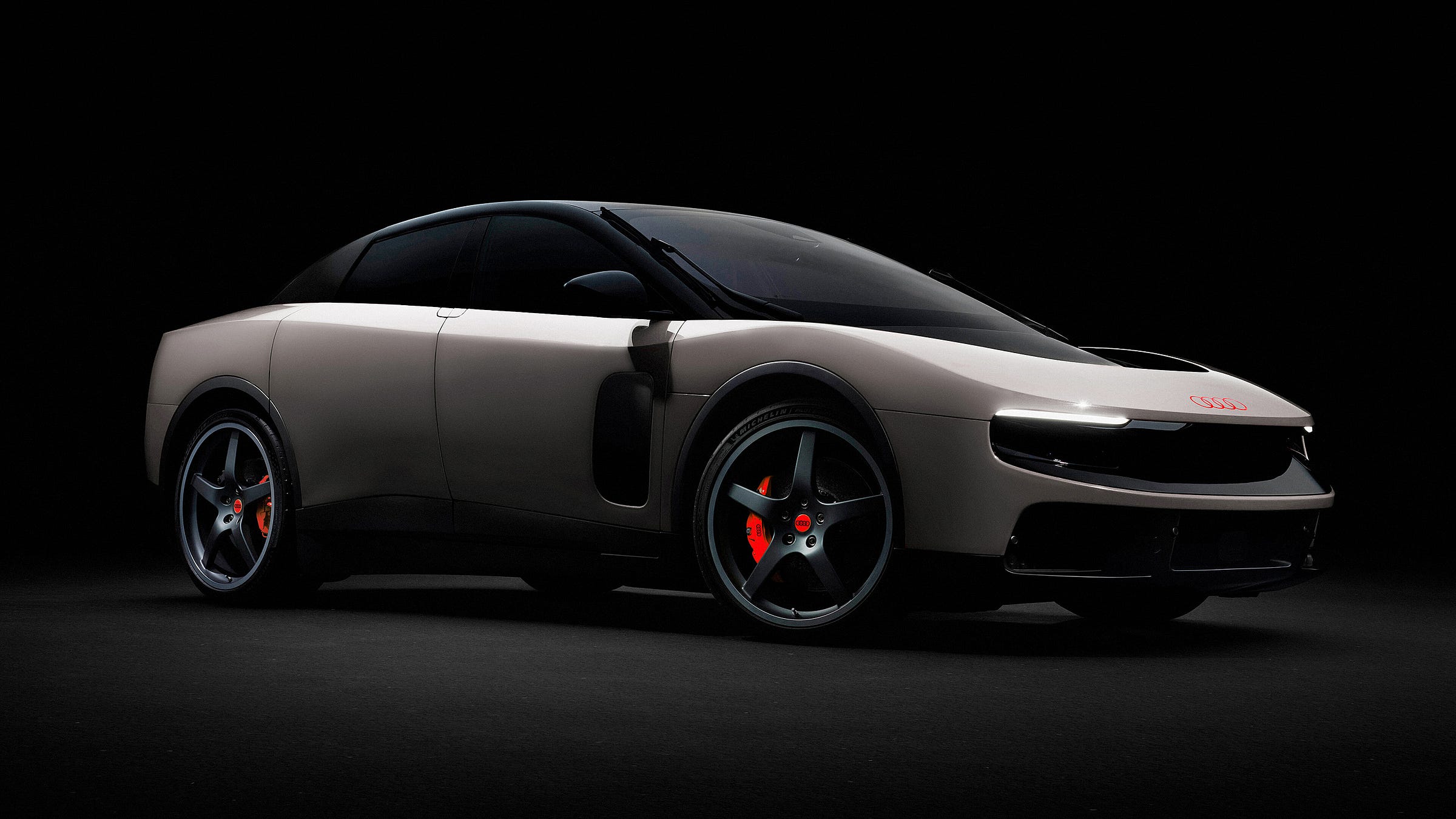A three-quarter front view of the Ferrari Luce in a dark studio setting, retouched to replace Ferrari branding with Audi's four-ring logo on the hood. The car is finished in a matte warm grey with a black roof, black five-spoke alloy wheels, and red brake calipers bearing the Audi logo.