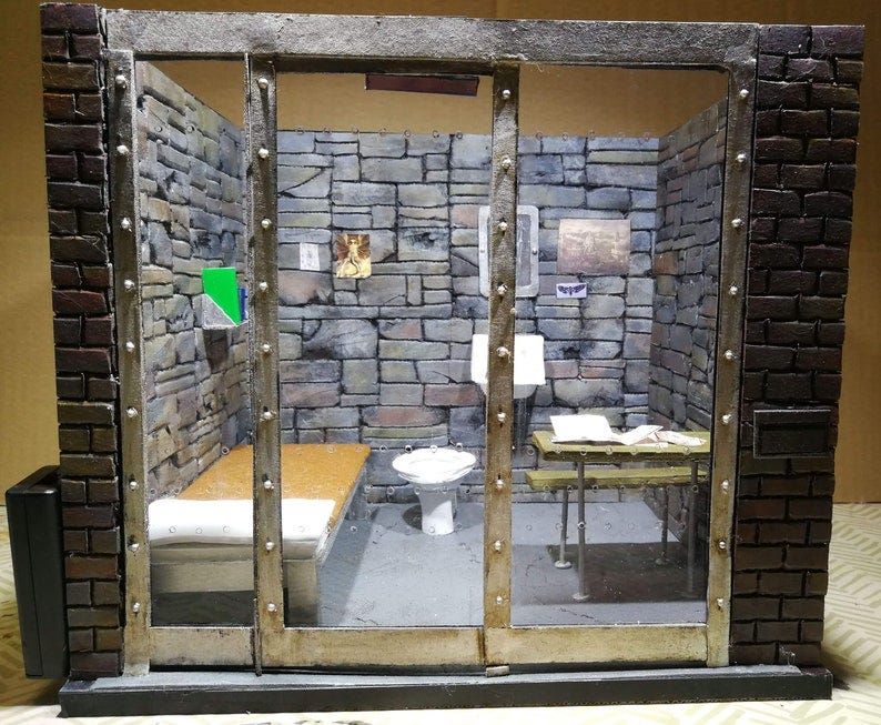 May include: A miniature model of a prison cell with a brick wall, a metal bed, a toilet, and a table. The cell is made of cardboard and has a glass front. The cell is lit from above and the walls are painted a dark gray color.