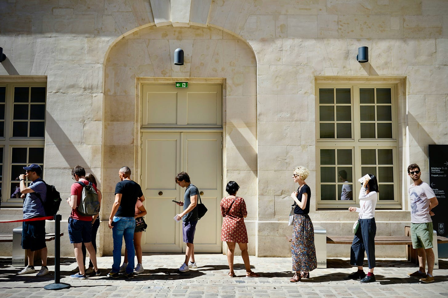 Image of people standingo outside in line on a sunny day, in front of an off-white building. 
