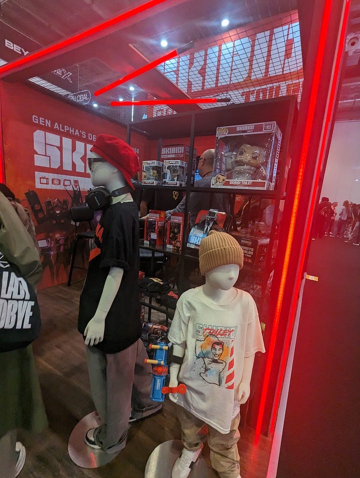 A trade show scene featuring two distinct exhibits: one showcasing an Oracle Red Bull Racing Formula 1 car displayed against a branded backdrop with sponsors such as Oracle, Red Bull, and Pirelli visible under bright convention hall lighting; and another booth promoting the Skibidi brand, where mannequins dressed in Gen Alpha–style streetwear stand beside shelves filled with Skibidi merchandise and Funko Pop figures, illuminated by striking red neon lights as visitors browse in the background.