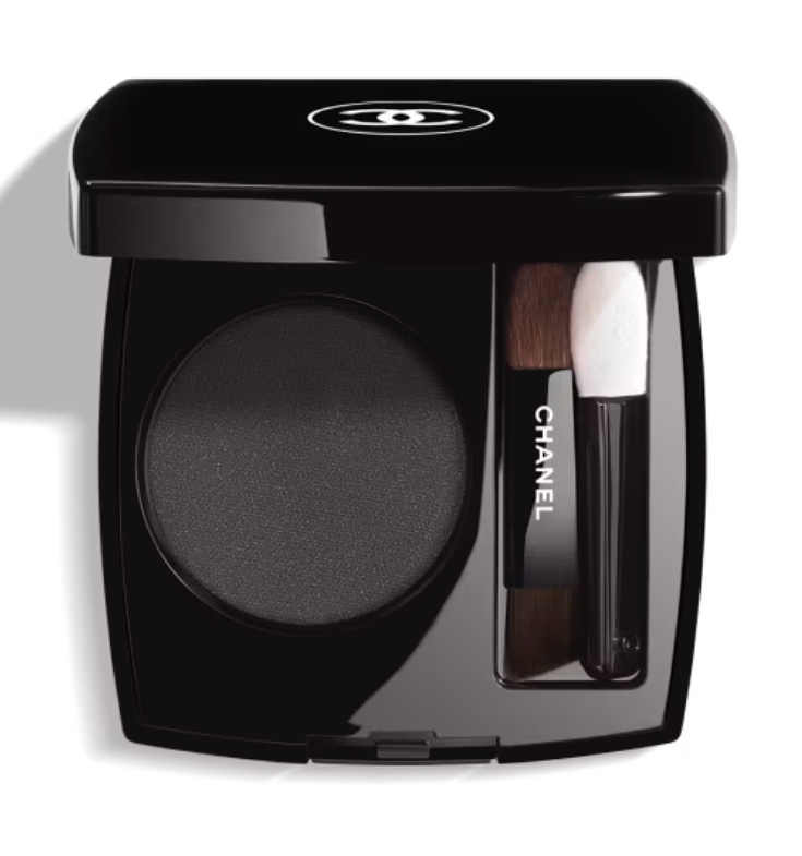 A top-down view of a classic black Chanel makeup compact with the white double-C logo on the lid; it is open to reveal a deep, matte charcoal-black powder eyeshadow with two mini applicators—a brush and a sponge tip.