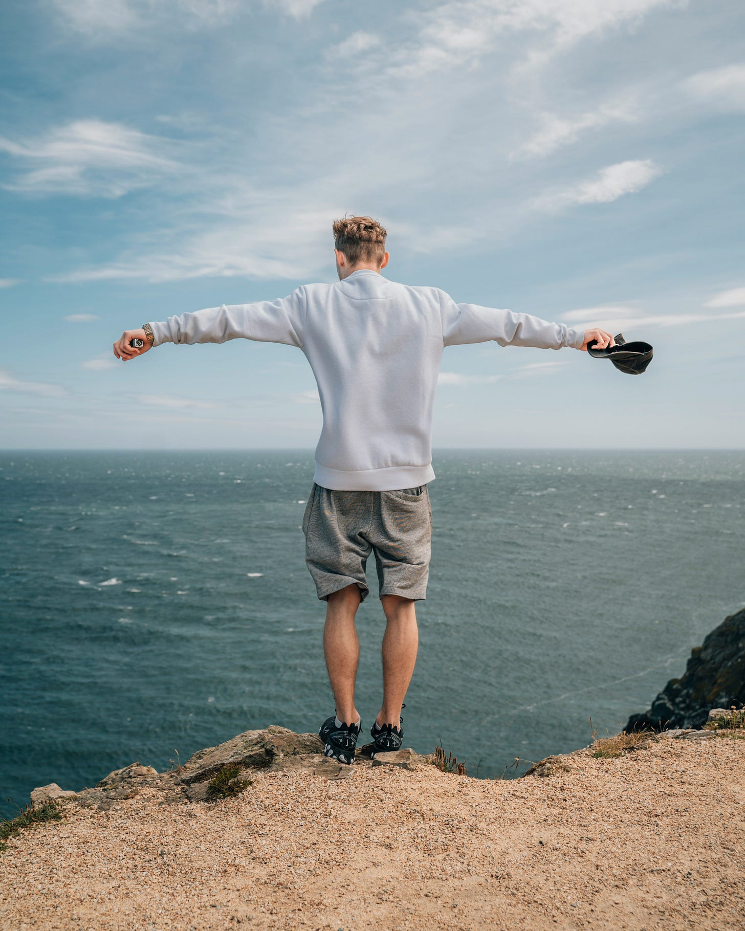 A person, seen from behind, perched on a cliff overlooking a blue body of water under a blue sky with whispy white clouds.