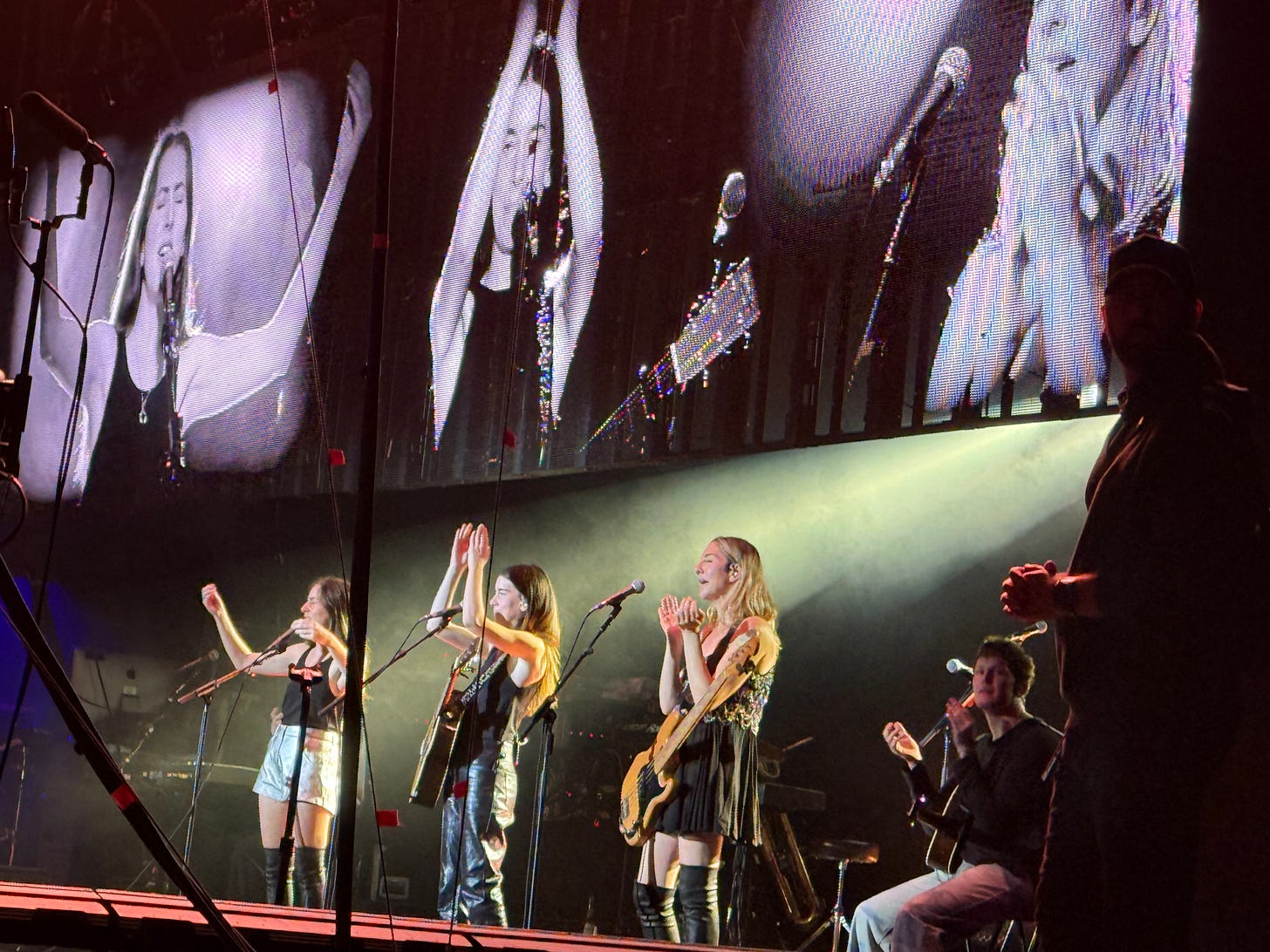 A group of female musicians on stage, smiling and applauding the audience. Large screens display black and white close-ups of their expressions. The atmosphere is energetic and celebratory.