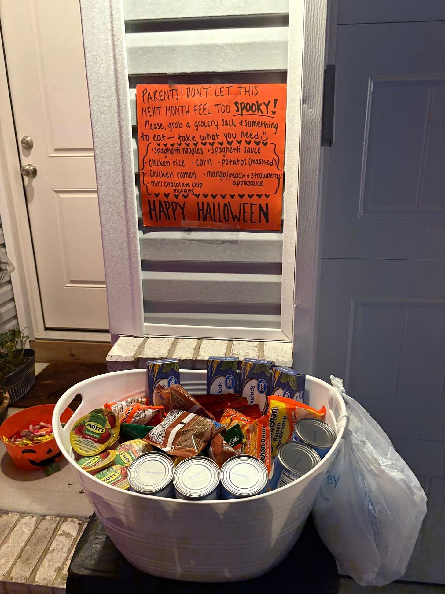 A front porch display with a large bowl filled with canned goods, ramen, and snacks under a handwritten orange Halloween sign inviting neighbors to take free food a community mutual aid project helping families during tough times.