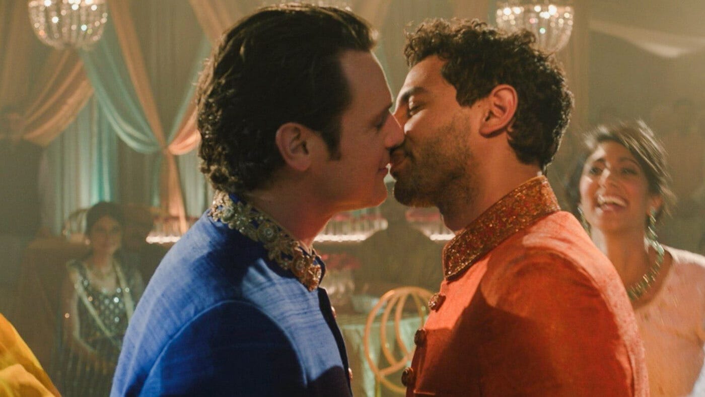 Naveen and Jay kiss in colorful sherwanis at a wedding celebration, surrounded by chandeliers and dancing guests.